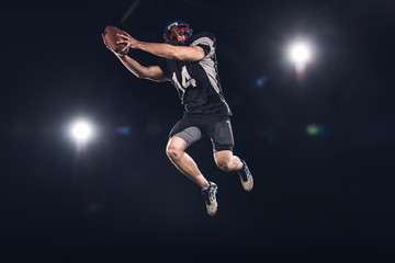 Fototapeta na wymiar bottom view of american football player jumping with ball under spotlights on black