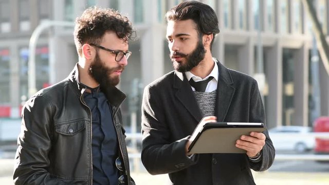 two  young  bearded  brown  hair  modern  businessman,  leaning  against  a  wall,  using  tablet  talking  and  discussing  -  technology,  business,  work  concept  