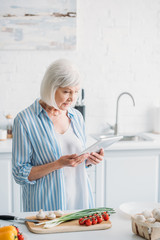 senior woman with digital tablet standing at counter with fresh vegetables in kitchen