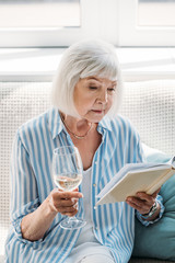 portrait of focused senior woman with glass of wine reading book on couch at home