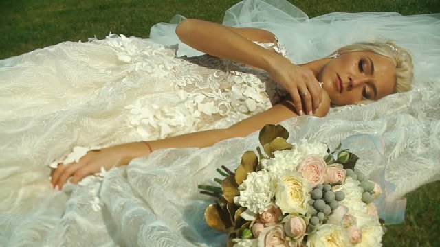 Beautiful tender blond bride in wedding dress is lying on the grass near the wedding bouquet.