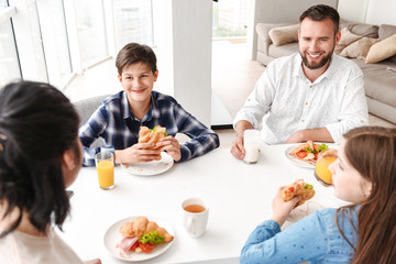 Image of happy parents with kids 8-10, sitting at table in bright kitchen, and having breakfast while eating croissant sandwiches