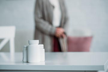selective focus of senior woman with walking stick standing in room with medicines on tabletop at home