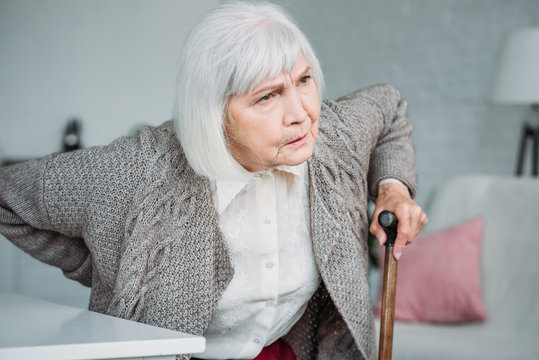 Portrait Of Grey Hair Lady With Back Ache And Wooden Walking Stick Sitting On Chair At Home