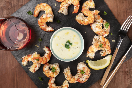 Overhead closeup photo of plate of cooked shrimps on a dark rustic background, with a sauce and a glass of wine, with copy space