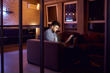 A man bearded freelancer with a laptop works in the evening in the room.