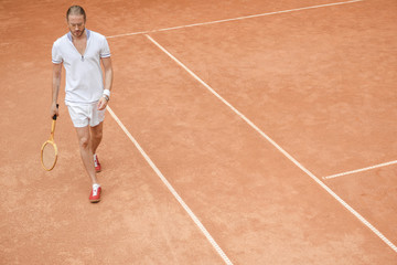 Fototapeta premium handsome sportsman with tennis racket walking on brown court