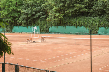 view of brown tennis court with net, chairs and trees