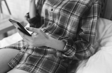 Beautiful girl is sitting on the white bed linen. Young woman in men's checkered shirt is searching in social network on white pillow. Morning with black smartphone in hands.