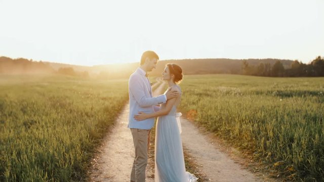 Bride and groom look at one another holding their hands and smiling during stroll on a countryside road in sunset light