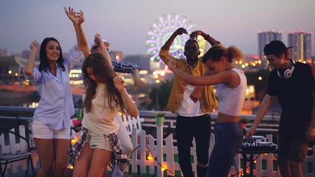 Slow motion of late rooftop party with joyful people dancing enjoying themselves and male deejay mixing modern music with professional equipment.
