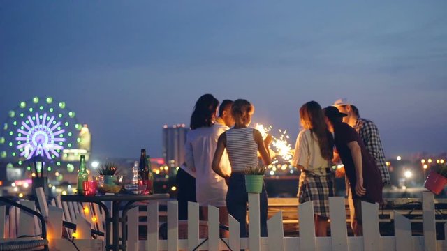 Slow Motion Of Happy People Friends Lighting Sparklers Then Raising Hands With Burning Bengal Lights At Night Party With Beautiful City In Background.