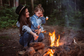 Mother and daughter fry marshmallow on  campfire in forest. Young family traveling active adventure...