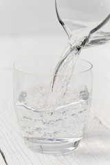 Pouring water from a glass jug into a glass. White wood background.