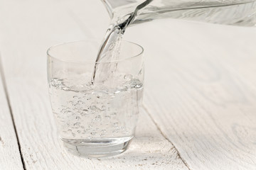 Pouring water from a glass jug into a glass. White wood background.