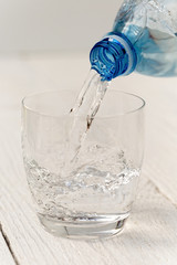 Pouring water from a plastic bottle into a glass. White wood background.