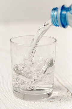 Pouring water from a plastic bottle into a glass. White wood background.