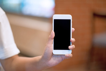 Mockup image of a woman holding and displaying a white mobile phone with a black screen with a blank screen on a table in a modern cafe. Ideas for working with communication tools.