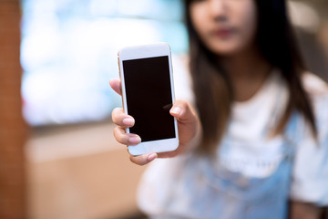 Mockup image of a woman holding and displaying a white mobile phone with a black screen with a blank screen on a table in a modern cafe. Ideas for working with communication tools.