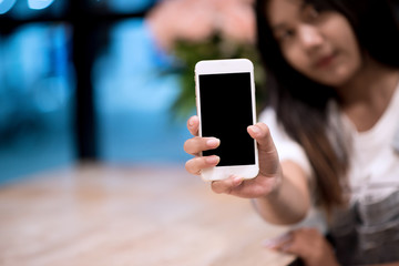 Mockup image of a woman holding and displaying a white mobile phone with a black screen with a blank screen on a table in a modern cafe. Ideas for working with communication tools.