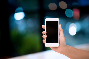Mockup image of a woman holding and displaying a white mobile phone with a black screen with a blank screen on a table in a modern cafe. Ideas for working with communication tools.