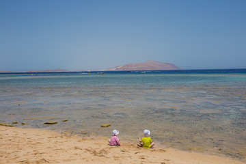 Two cute little toddler babies sit on sandy beach near blue sea water. Horizontal color image.