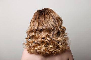Female hairstyle with long curls on the head of a blonde with a back view on a gray background.
