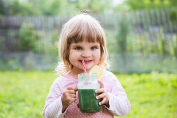 Cute little child drinks healthy green smoothie with straw in a jar mug against the background of greenery outdoor. 