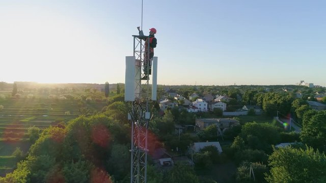 technician on top of cellular antenna show thumb up on background of blue sky and sunlight, aerial shot of telecommunication tower