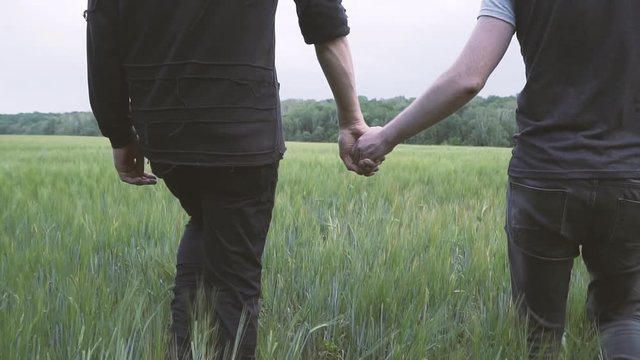 A couple of gay men walking on a green wheat field holding hands.