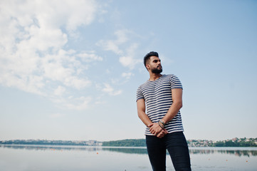 Handsome tall arabian beard man model at stripped shirt posed outdoor against lake and sky. Fashionable arab guy.