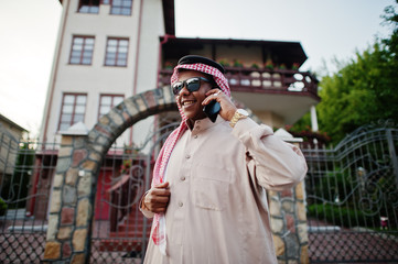 Rich Middle Eastern arab business man with sunglasses posed on street against mansion , speaking on mobile phone.