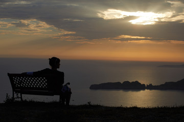 Fototapeta na wymiar Young woman sits on a bench on a sunset background over the sea and mountains