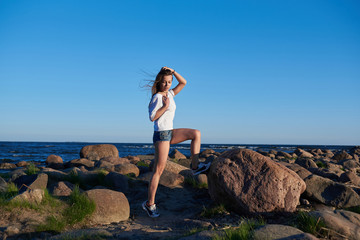 The girl stands on a large stone against the backdrop of rocks and the sea and enjoys the sunset
