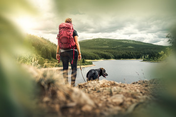 young woman with backpack and german shepherd dog puppy standing on mountain in front of forest and lake