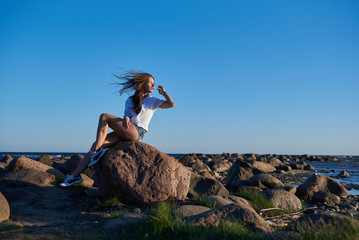 The girl sits on a large stone against the backdrop of rocks and the sea and enjoys the sunset