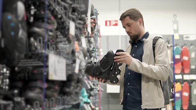 Young guy with a beard wants to buy brand new roller skates for skating in summer. Man wants to get fit doing cardio on roller skates. Sport shop.
