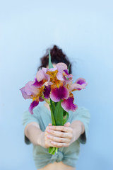 The girl in a green t-shirt with arms outstretched holds a bouquet of bright beautiful irises, covers her face, against the blue wall