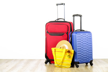 Red textile suitcase & blue hard shell luggage, extended telescopic handle, straw hat, yellow beach bag, mirrored sunglasses, white wall background. Couples retreat trip concept. Close up, copy space.