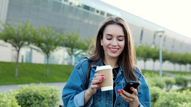 Close up view of Pretty Young Woman Walking through the Park. Using her Mobile Phone. Green Park near the Modern Building. Woman is Smiling with Pleasure. Stylish Outfit.