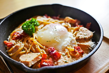 Sizzling hot and creamy Spaghetti Chicken Carbonara topped with Onsen Egg in a black cast iron pan on a wooden table. Selective focus.