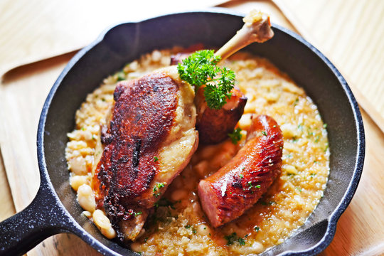 Sizzling Hot, Rich, Slow-cooked Trio Cassoulet Of Duck Confit, Pork Belly And Smoked Sausage On A Bed Of White Beans In A Black Cast Iron Pan On A Wooden Table.