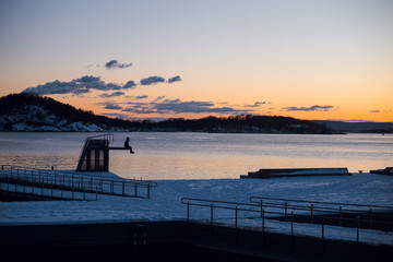 Obraz premium Young hiker man sitting on a pier and looking on the calm lake and mountains on beautiful pink and orange sunset. Twilight. Norway