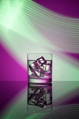 A glass with a drink and ice cubes on a magenta background with green lights.