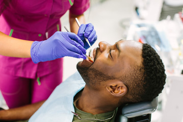 Close-up portrait of the african young man patient examining by female dentist with tools in dental clinic. Doctor doing dental treatment on man's teeth in the dentists chair.