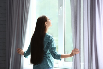 Young woman opening curtains in room near big beautiful window