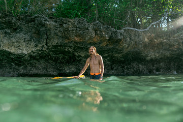 surface level of shirtless smiling male surfer standing in ocean with surfing board in Bali, Indonesia