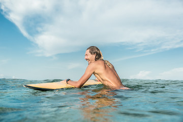 side view of smiling shirtless male surfer swimming with surfing board in ocean at Nusa Dua Beach, Bali, Indonesia