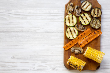 Grilled vegetables on rustic wooden board on a white wooden background, top view. From above, flat lay, overhead. Copy space and text area.