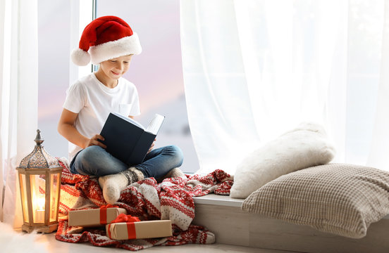 Cute Little Child In Santa Hat Reading Christmas Story While Sitting On Windowsill At Home